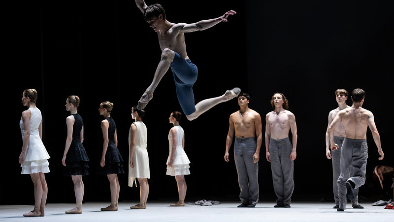 Ballerino der Hamburger Staatsoper, schwebt in einer ausdrucksstarken Pose in der Luft, während andere Tänzer im Hinterdrund stehen. Alle Kleider wurden von Akris entworfen.